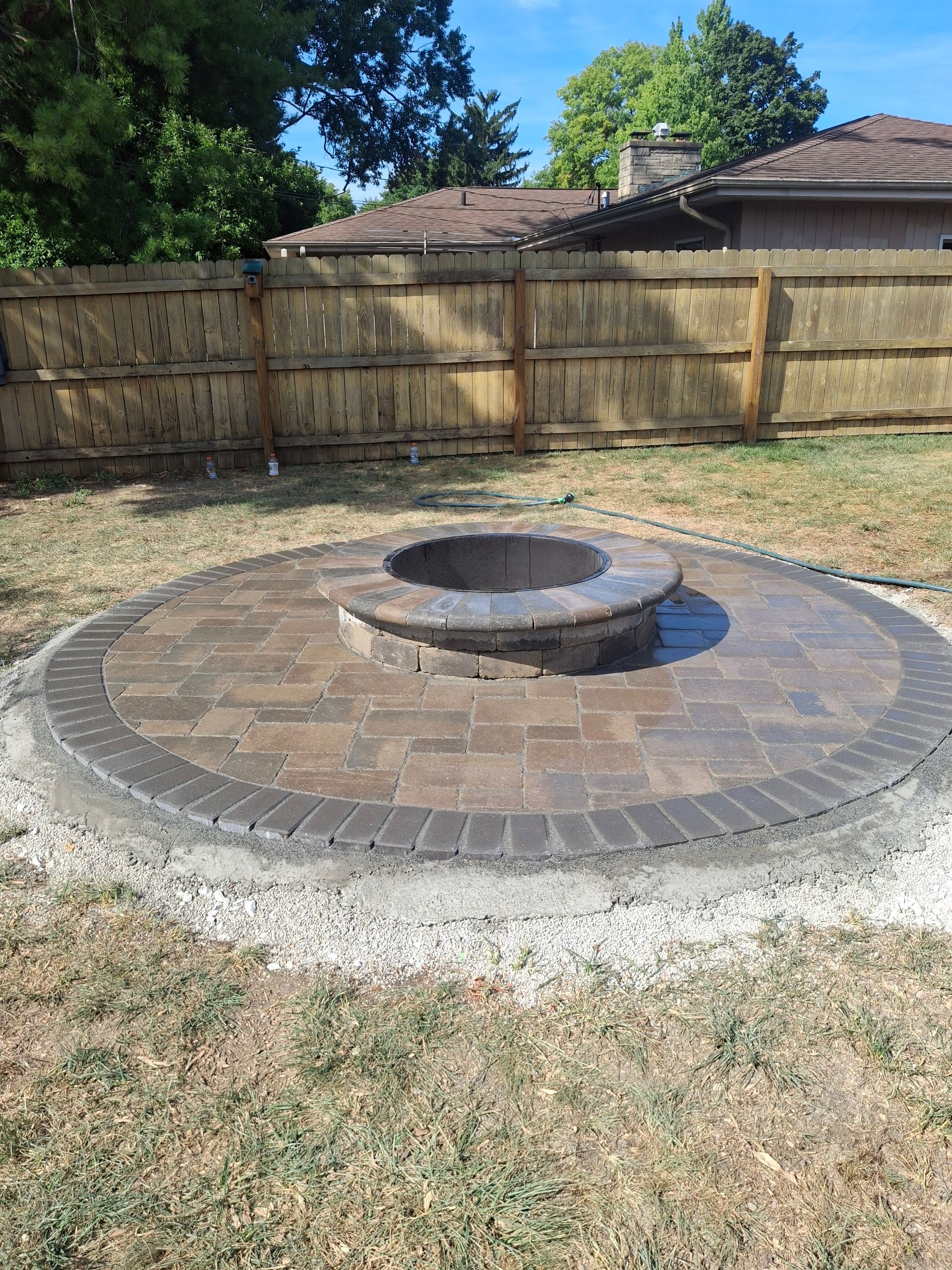Circular brick patio with fire pit, surrounded by a wooden fence and grass.
