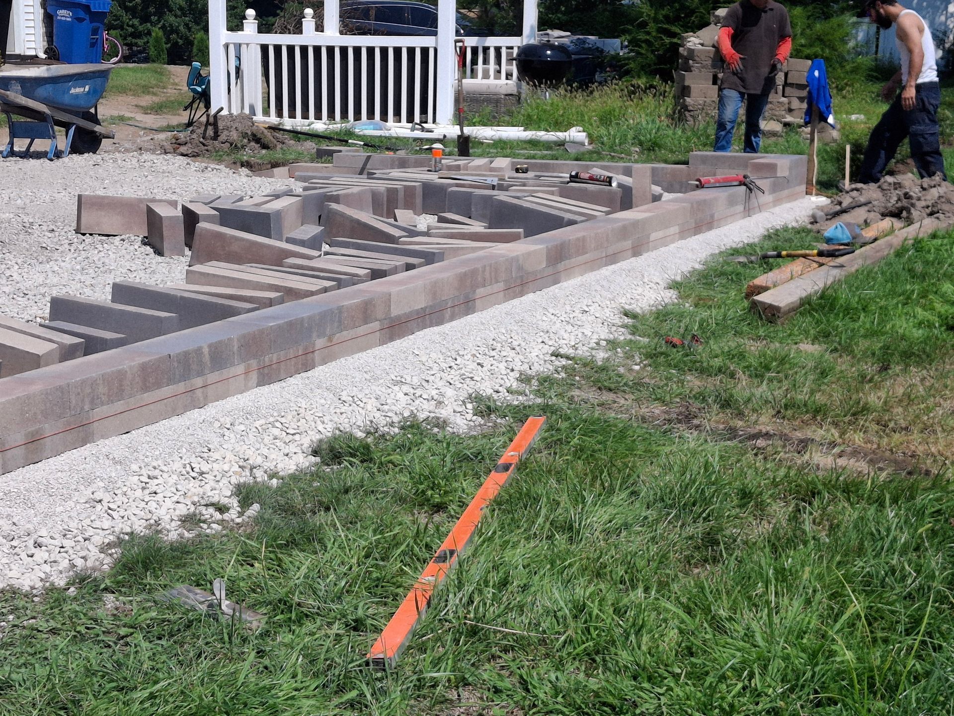 Construction site: Workers building a retaining wall with paving stones. White gravel base, green grass, and a white gazebo in the background.
