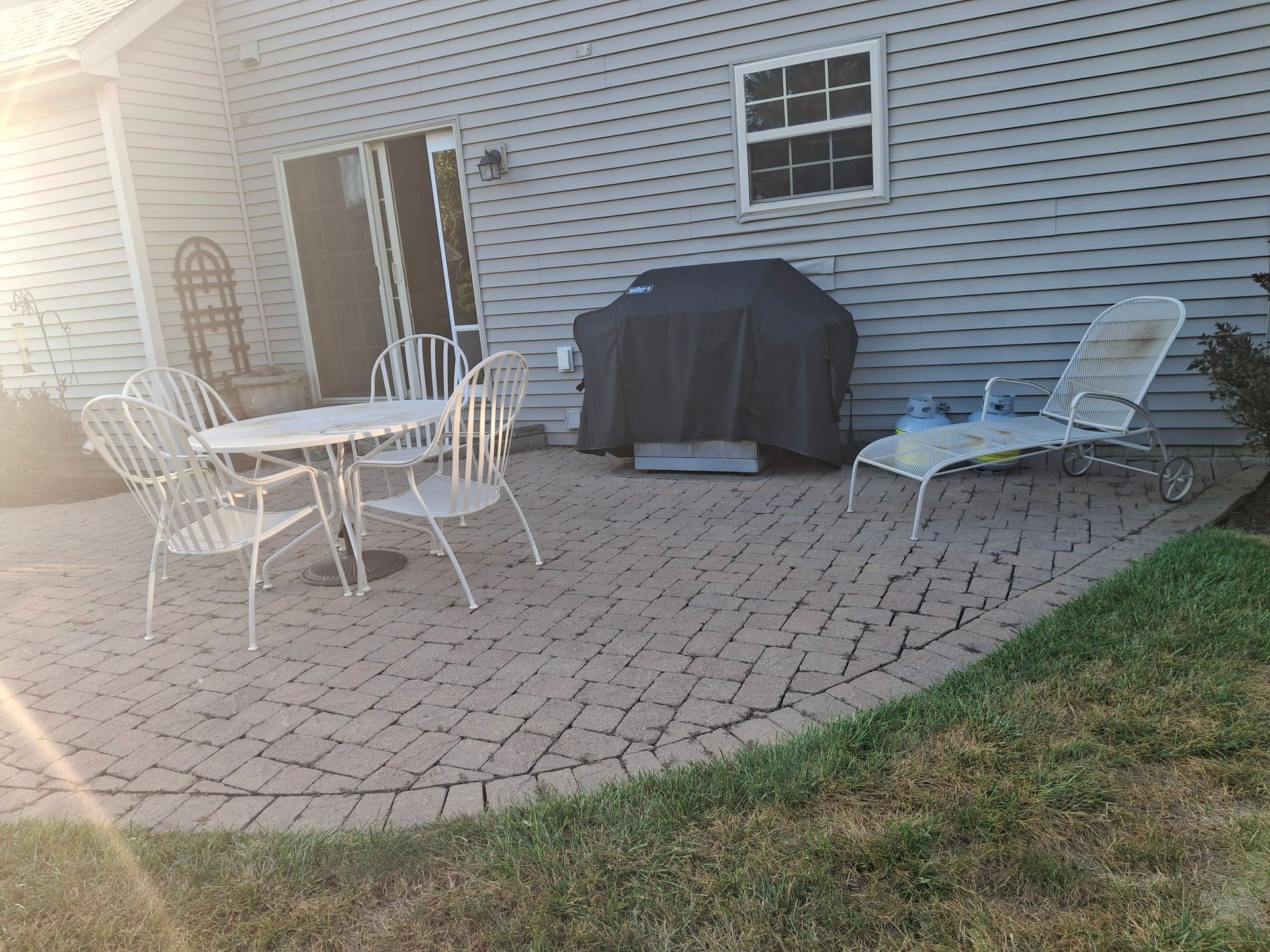 A patio with a table and chairs, a grill, and a chaise lounge next to a house with green grass.