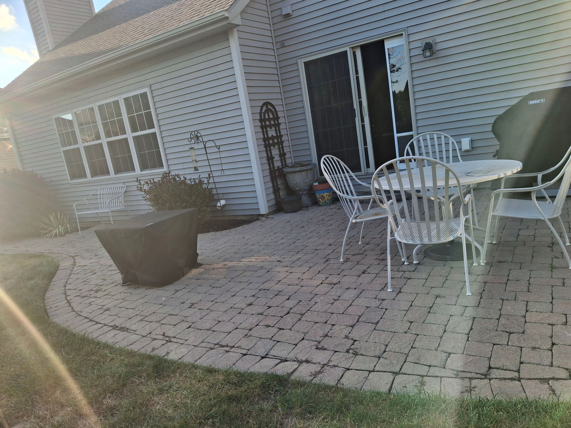 Brick patio with white chairs and table, next to a house with a sliding glass door.