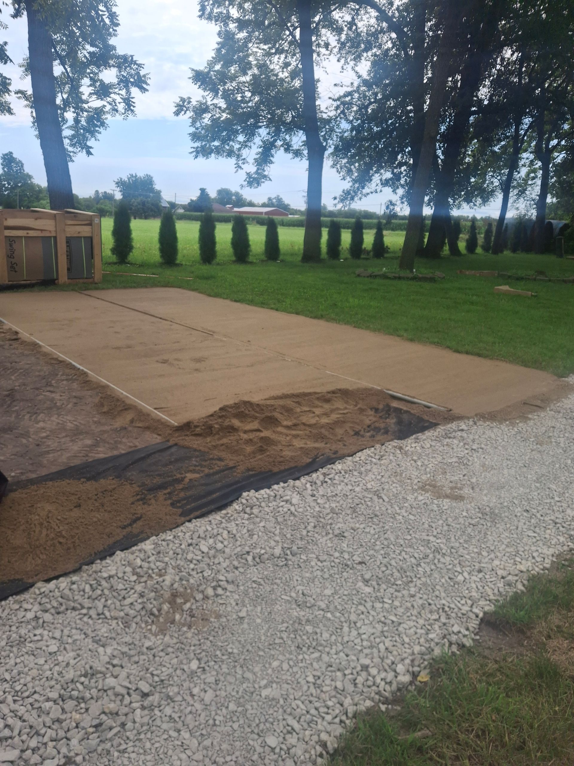 Construction site with gravel path, sand, and grass. Wooden crate and line of trees in the background.