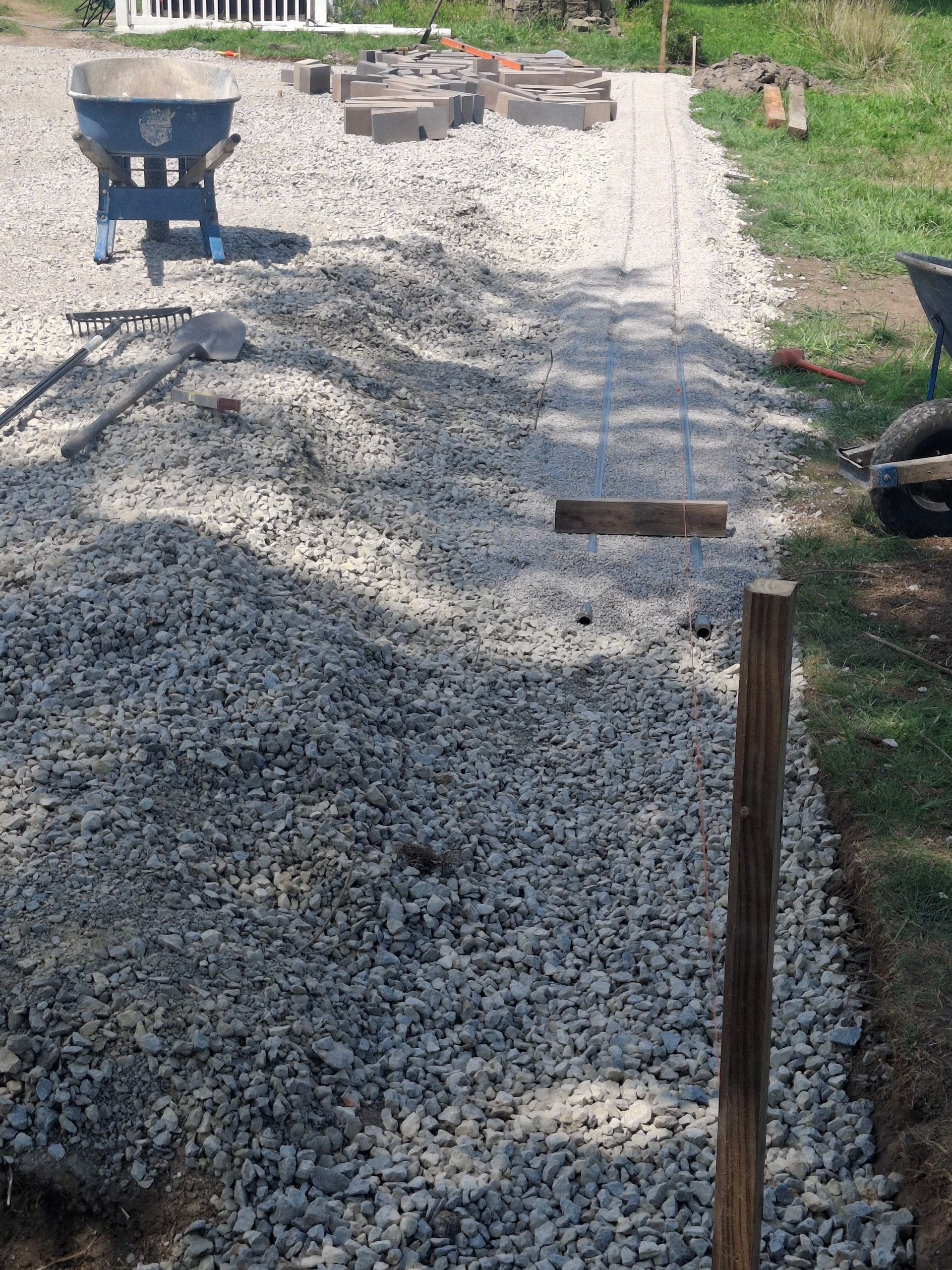 A gravel path under construction with stone pavers, bordered by grass. A wheelbarrow and building supplies are nearby.