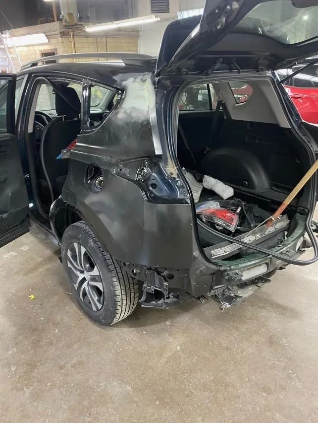 Black SUV with rear damage, in a repair shop, with the hatch and door open.