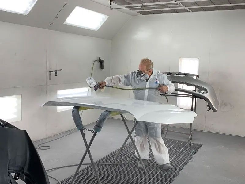 Person in protective gear spray-painting a car hood white in a paint booth.