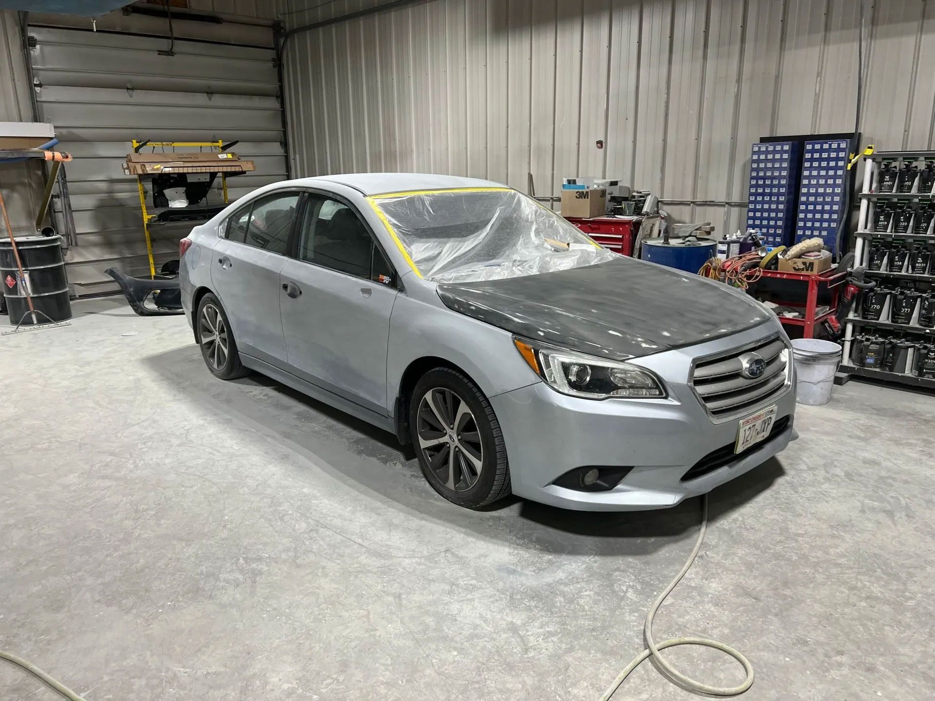 Silver car in a repair shop with the hood and roof covered in plastic, ready for paint.