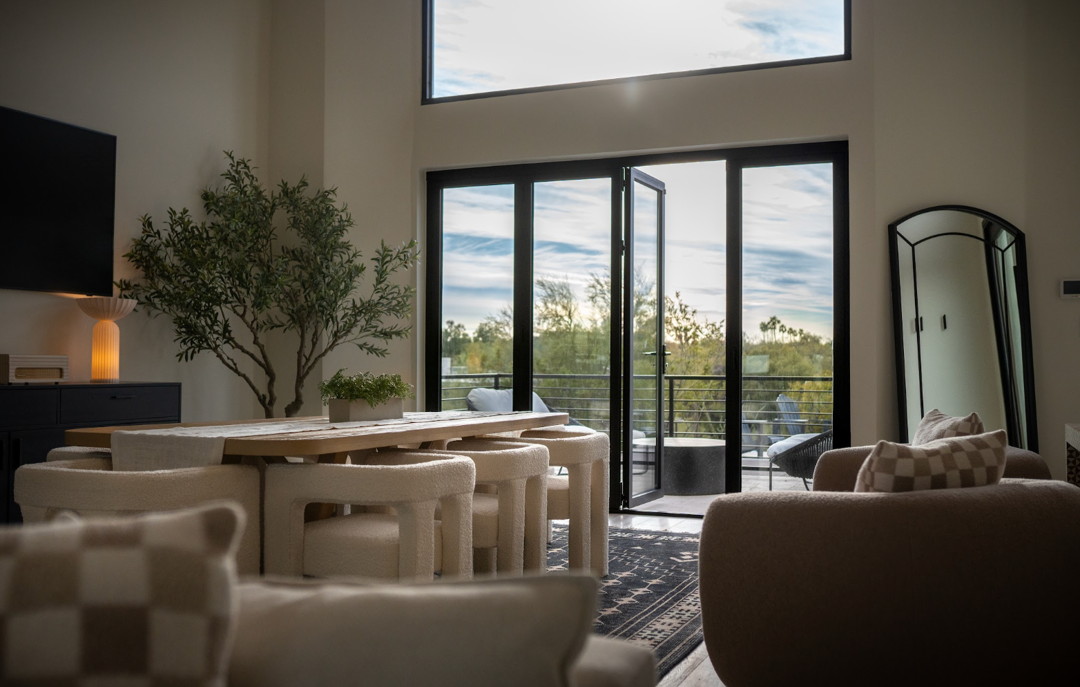 Modern living room with large glass doors, dining table, sofa, and indoor tree overlooking greenery.