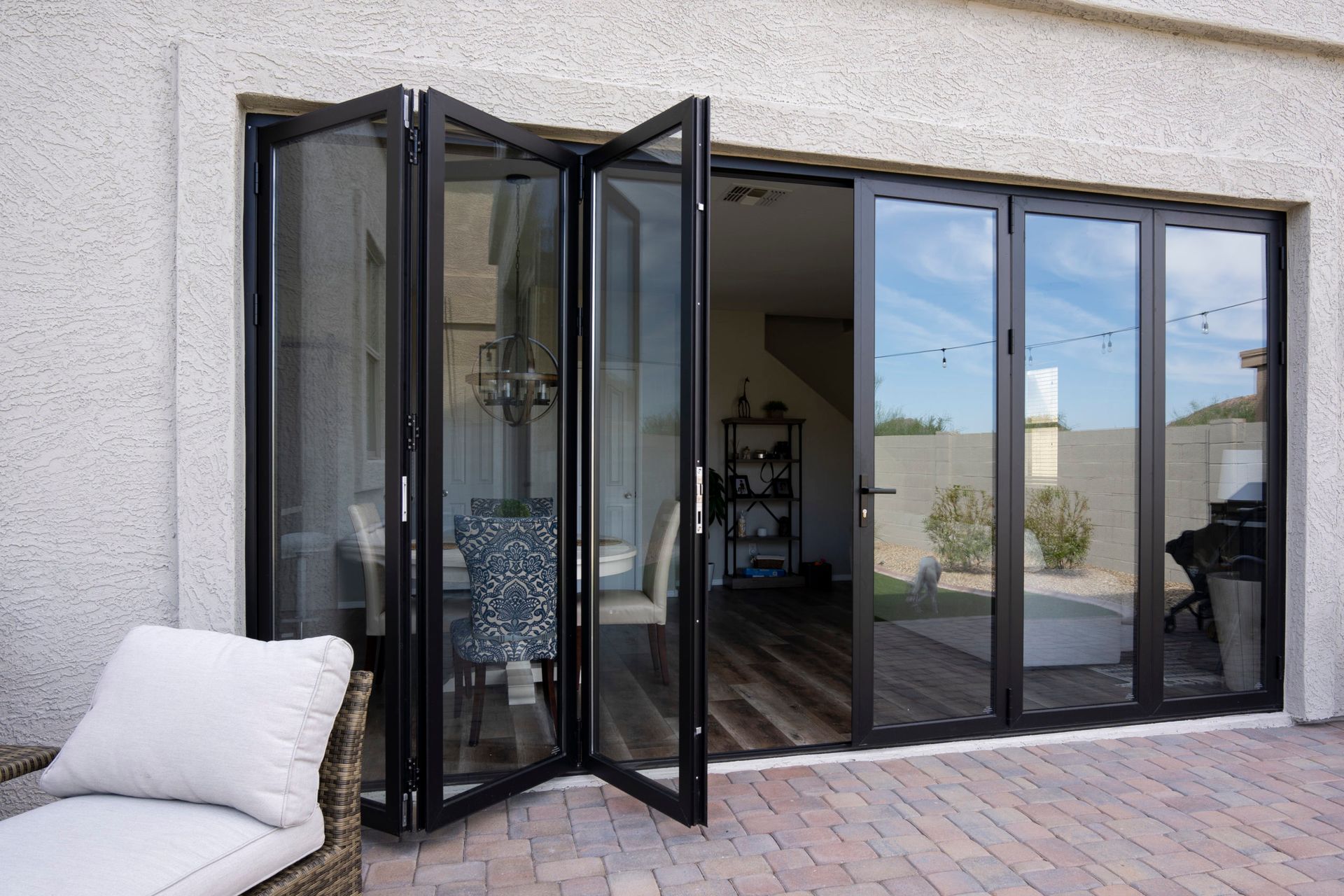 Folding glass doors open onto a patio, revealing a home's interior. Black frames, neutral exterior, and patio furniture.