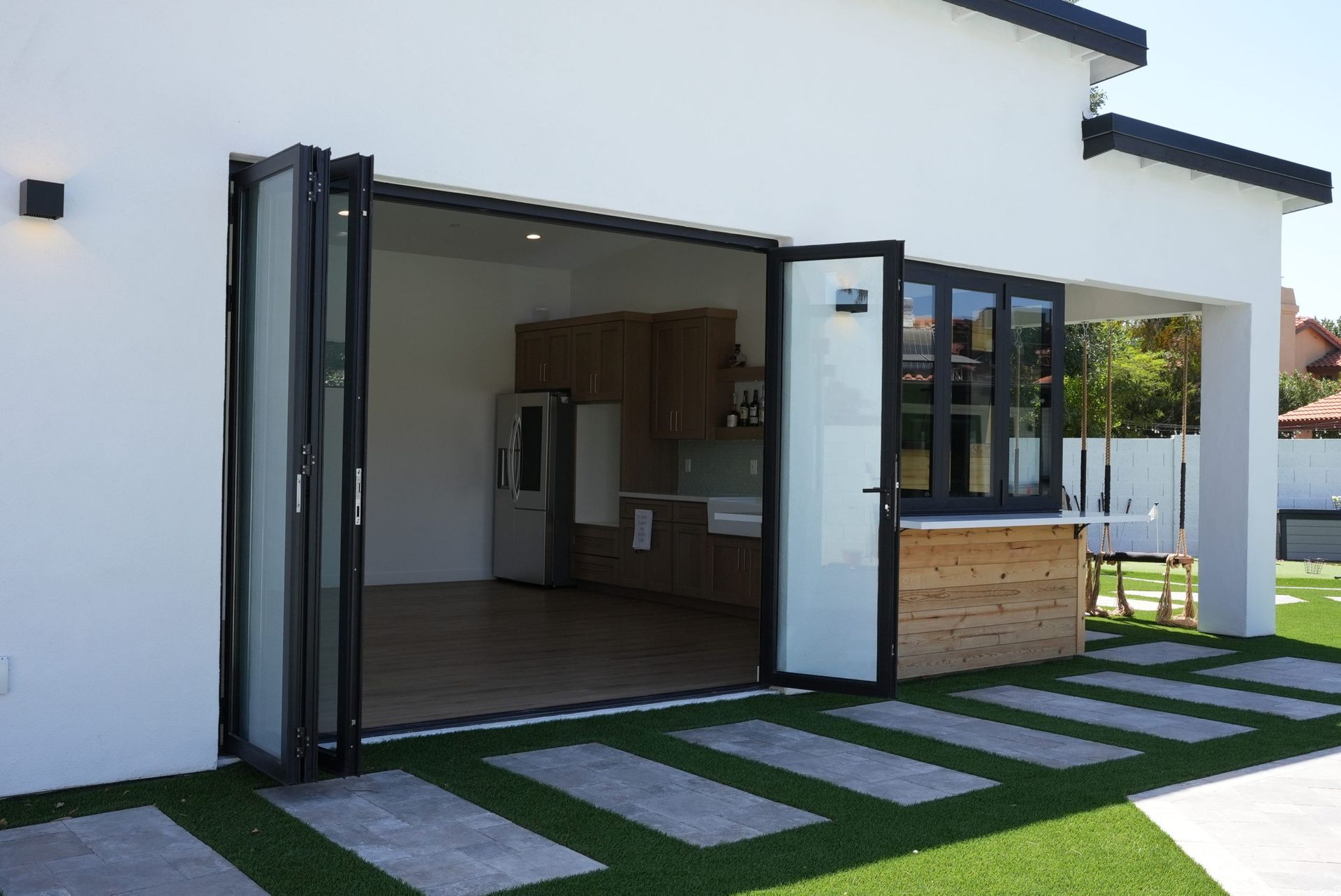 Exterior of a house with open folding doors to a kitchen and outdoor bar. Grass and stone pathway.