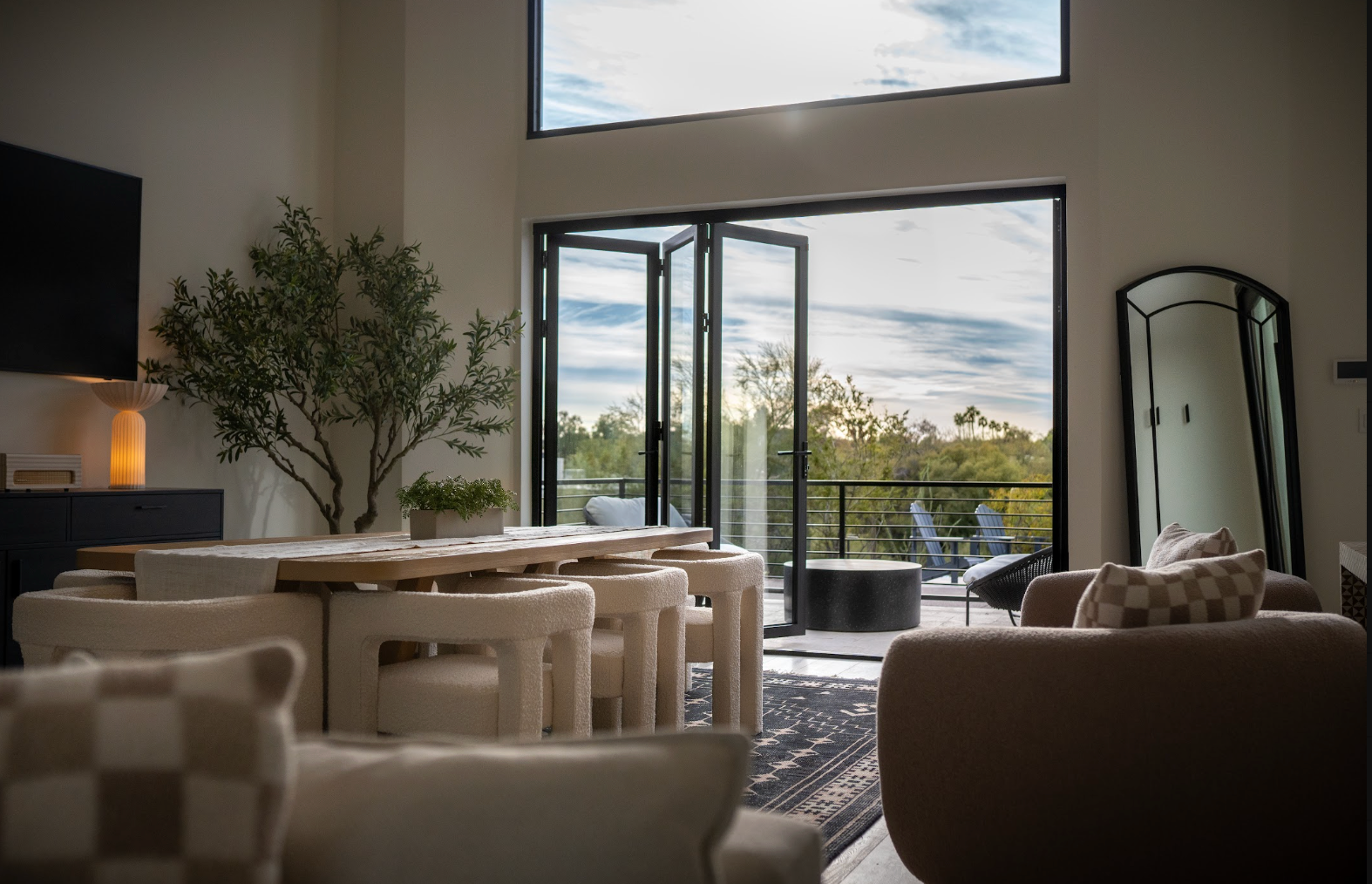 Modern living room with open glass doors to a patio and sunset view, featuring neutral furniture and a potted tree