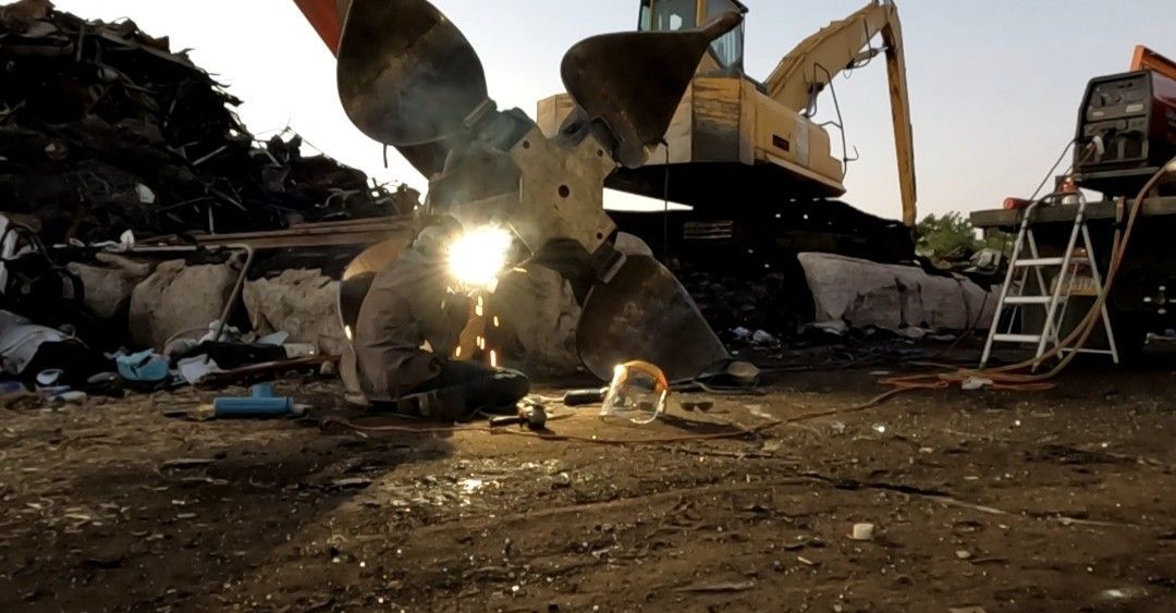 A large metal claw machine cutting through scrap metal at a yard, creating bright sparks in the center.