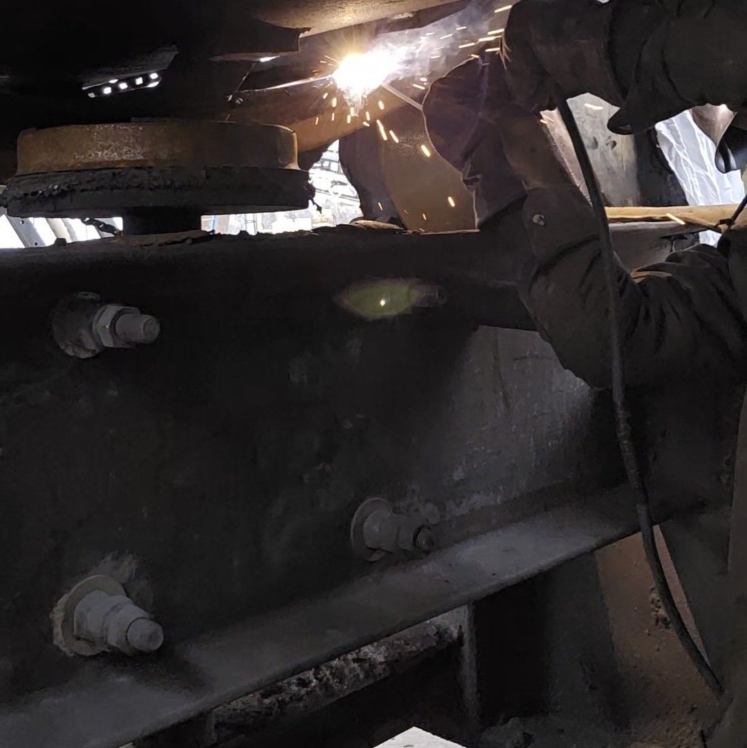 A person wearing protective gloves uses a welding torch on a heavy metal frame, creating bright sparks.