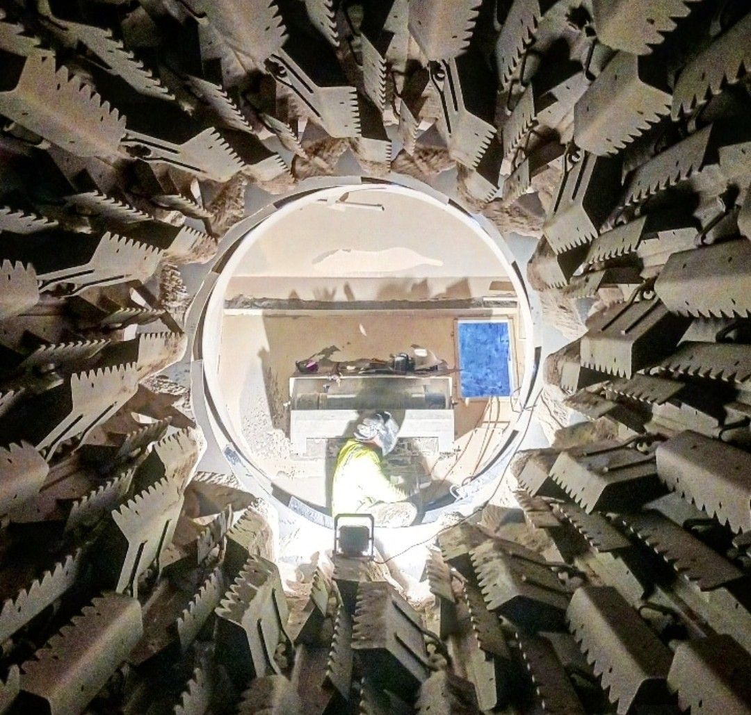 A worker in a high-visibility vest stands inside a tunnel-boring machine, surrounded by large, circular metal cutting teeth.