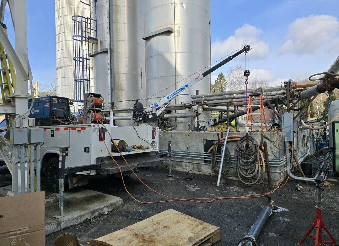 A service truck with a boom crane parked in an industrial yard near large metal storage silos under a blue sky.
