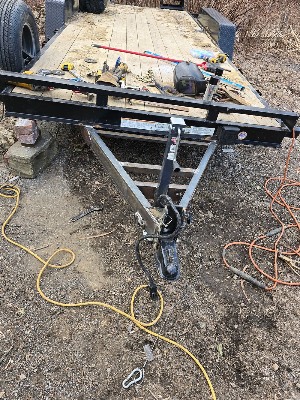 Black utility trailer tongue on gravel with tools and extension cords nearby.