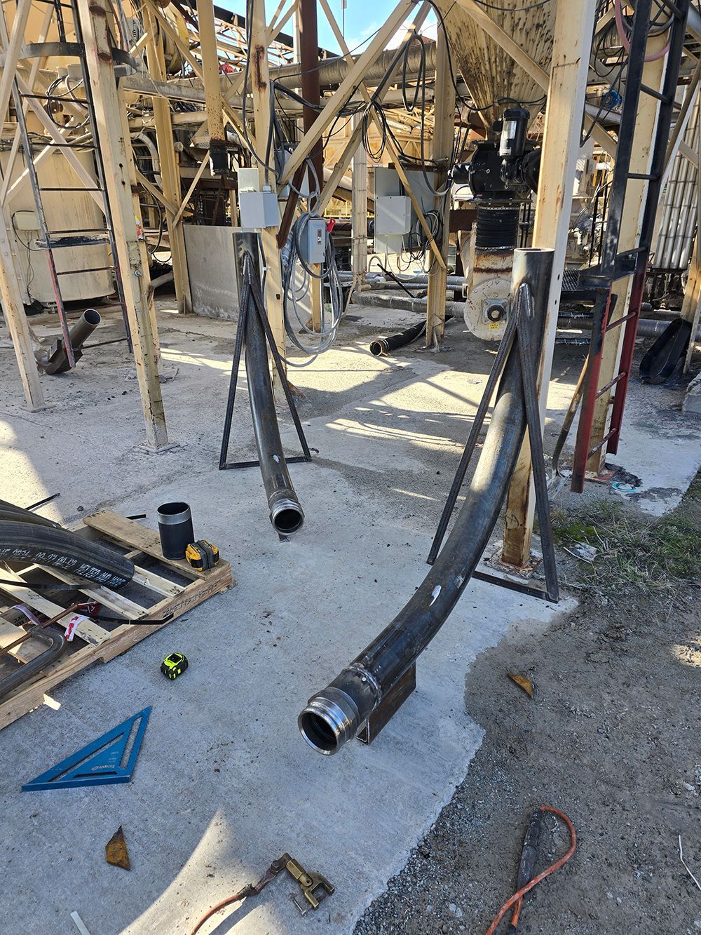 Collapsed black pipes hanging over a concrete industrial worksite, with scattered debris and tools