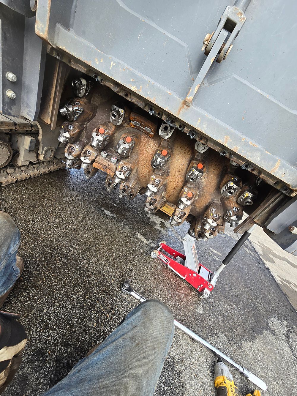 Exposed water meter and pipe manifold under a metal panel, with wet gravel and tools on the ground