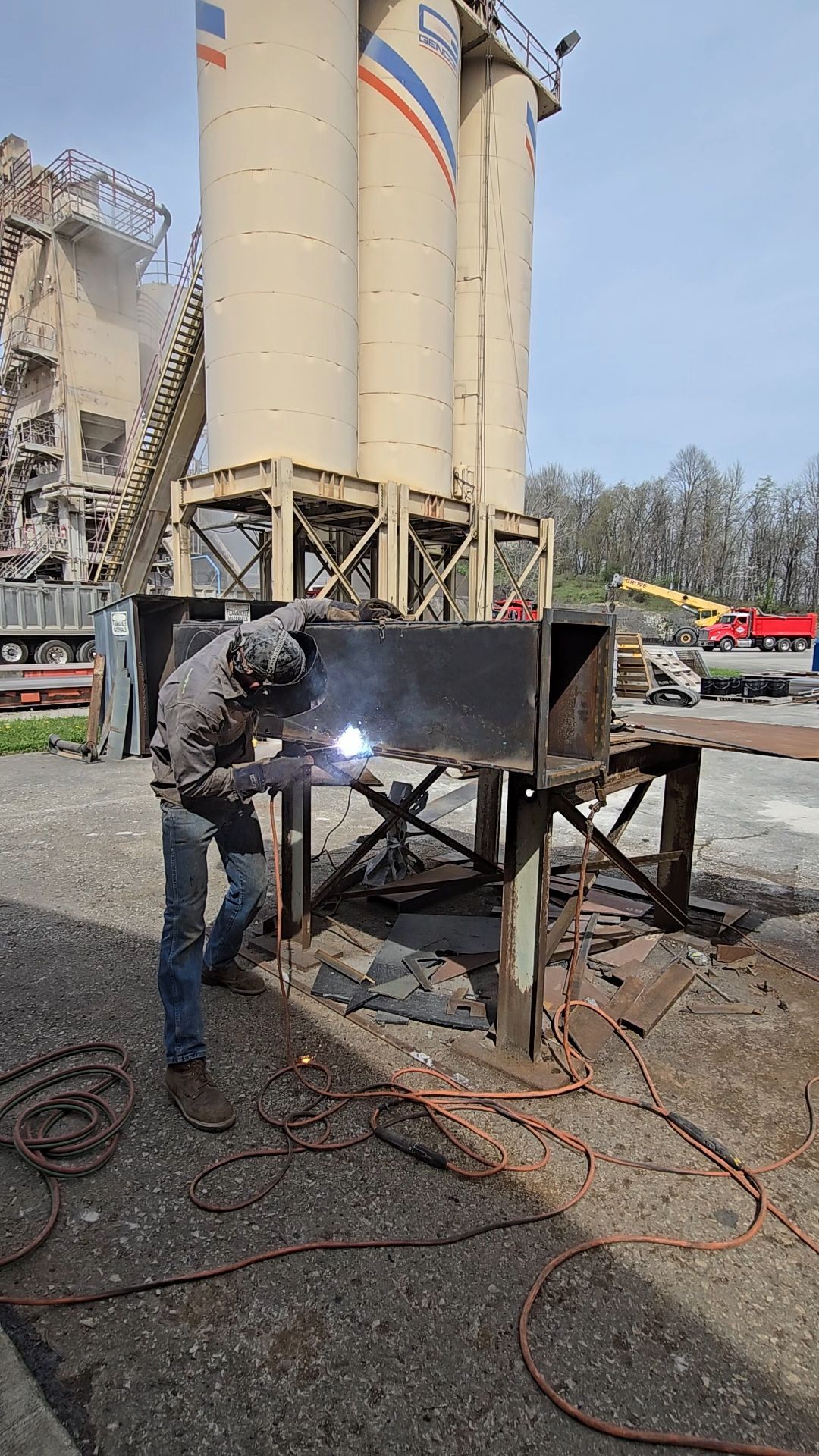 Worker welding a metal hopper outdoors beside large silos and construction equipment