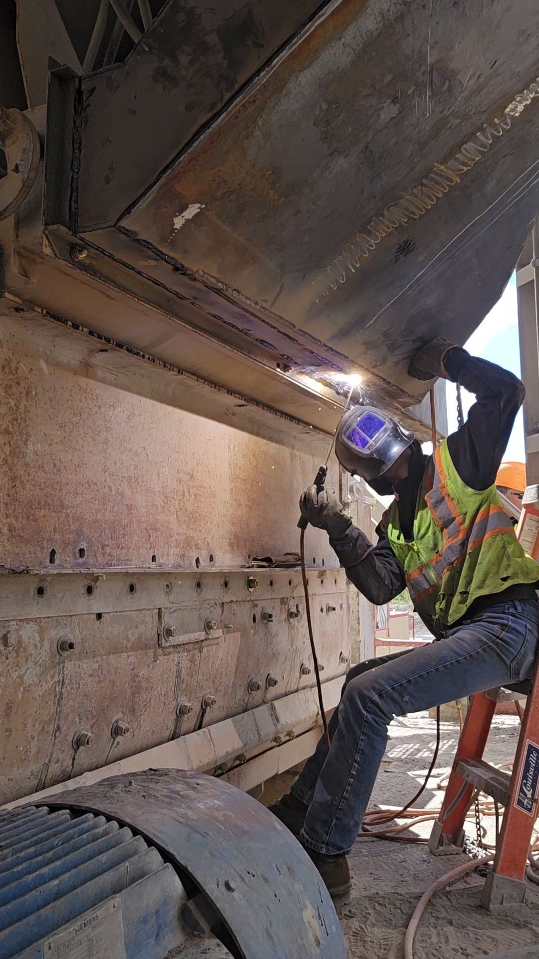 Worker in high-vis gear welding under a large metal structure, sparks visible, with a ladder nearby