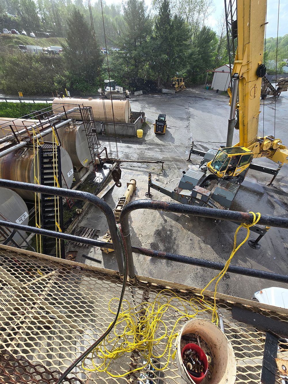 Industrial worksite with a yellow crane, pipes, and workers on a wet gravel yard