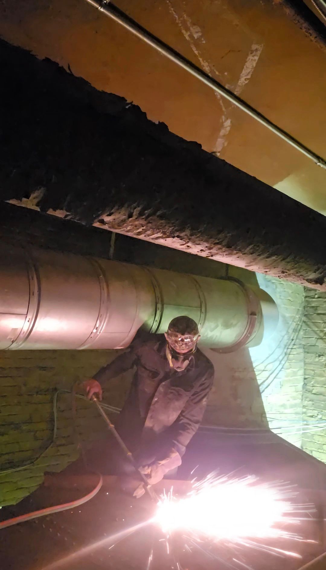 A worker welding under large pipes in a dim industrial setting, sparks flying brightly.