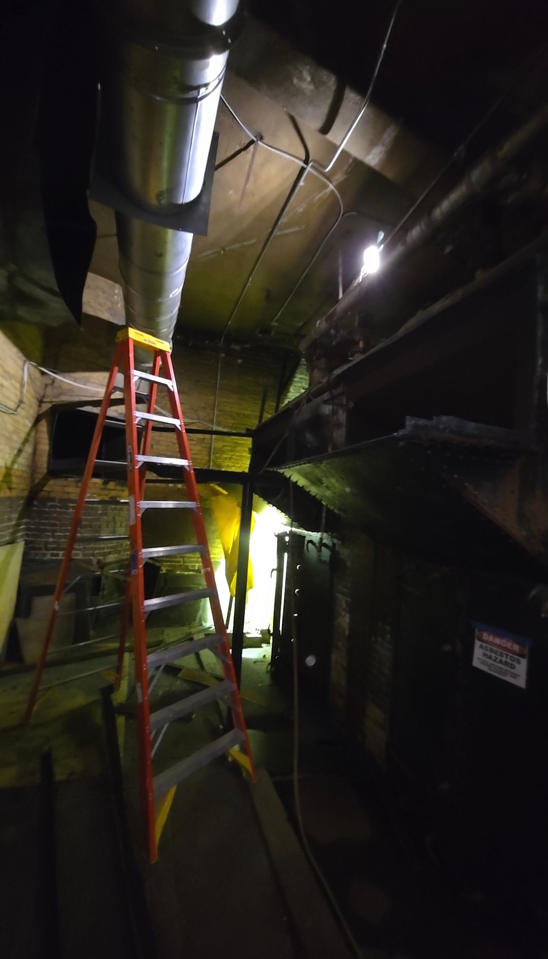 Red ladder in a dim industrial hallway with exposed ductwork and pipes overhead.