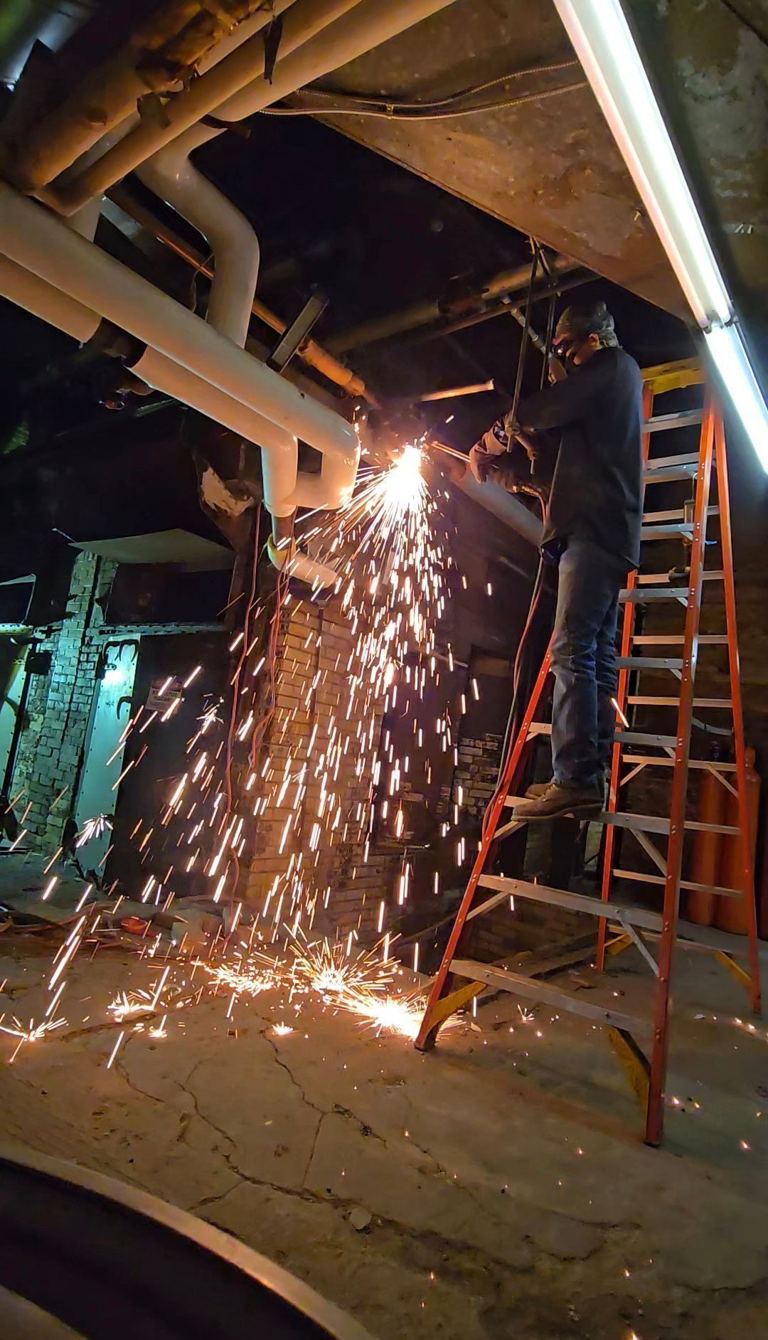 Worker cutting metal overhead, sparks showering down beside a ladder in an industrial setting