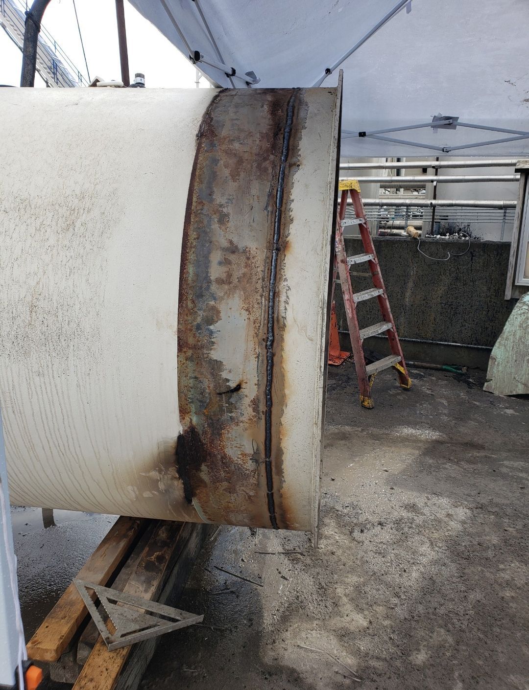 Rusted metal band on a large white pipe under a tent, with a red ladder and industrial equipment nearby