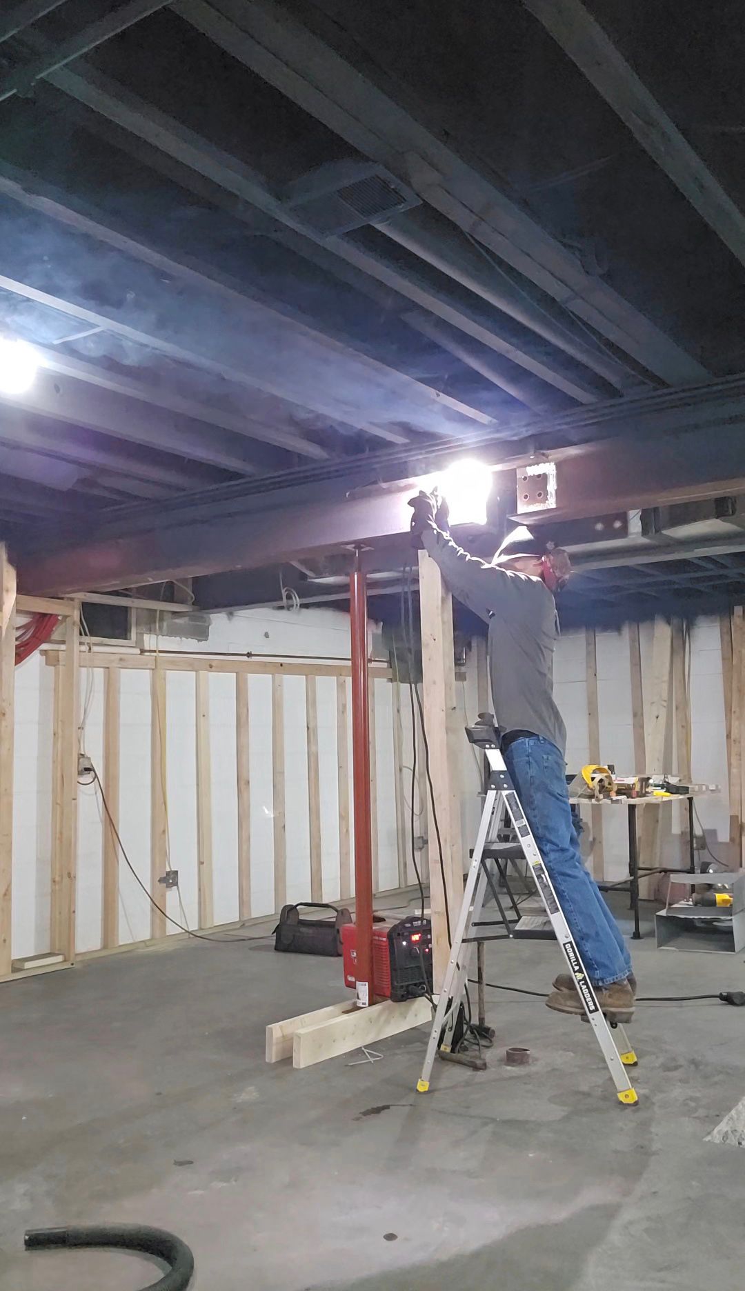 Worker on a ladder installing ceiling lights in an unfinished basement.