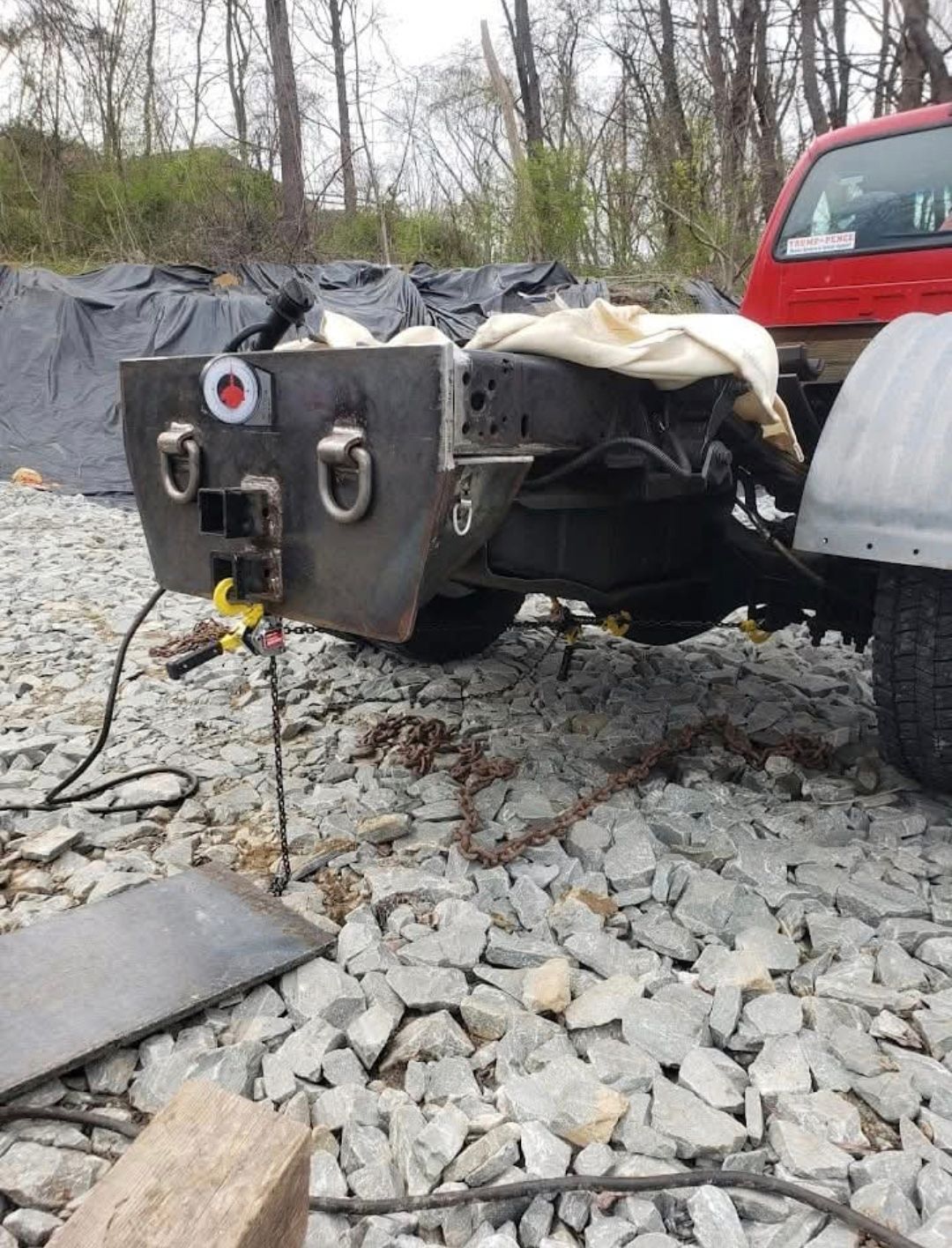 Overturned black trailer on gravel beside a red truck in a wooded area