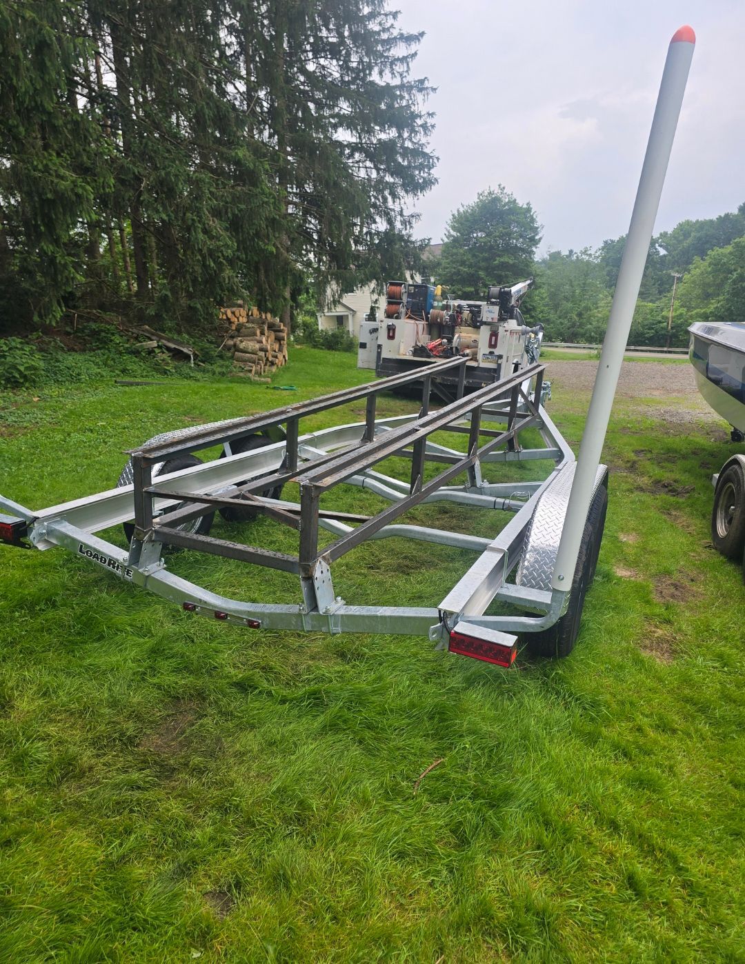 Boat trailer on grass near water, with a long white pole and work vehicle in the background