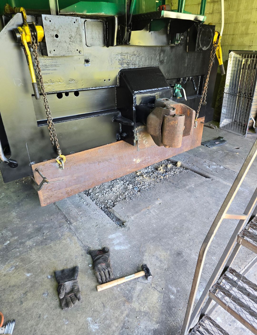 Industrial machine with heavy metal parts and chains in a workshop, with gloves and a hammer on the floor.
