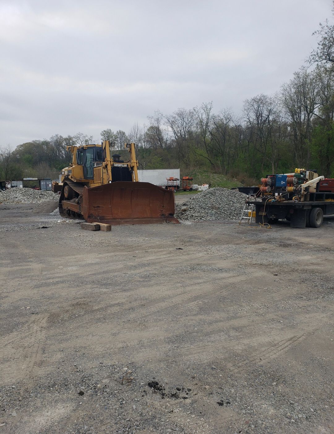Bulldozer and work trailer in a gravel lot with piles of rock and trees in the background
