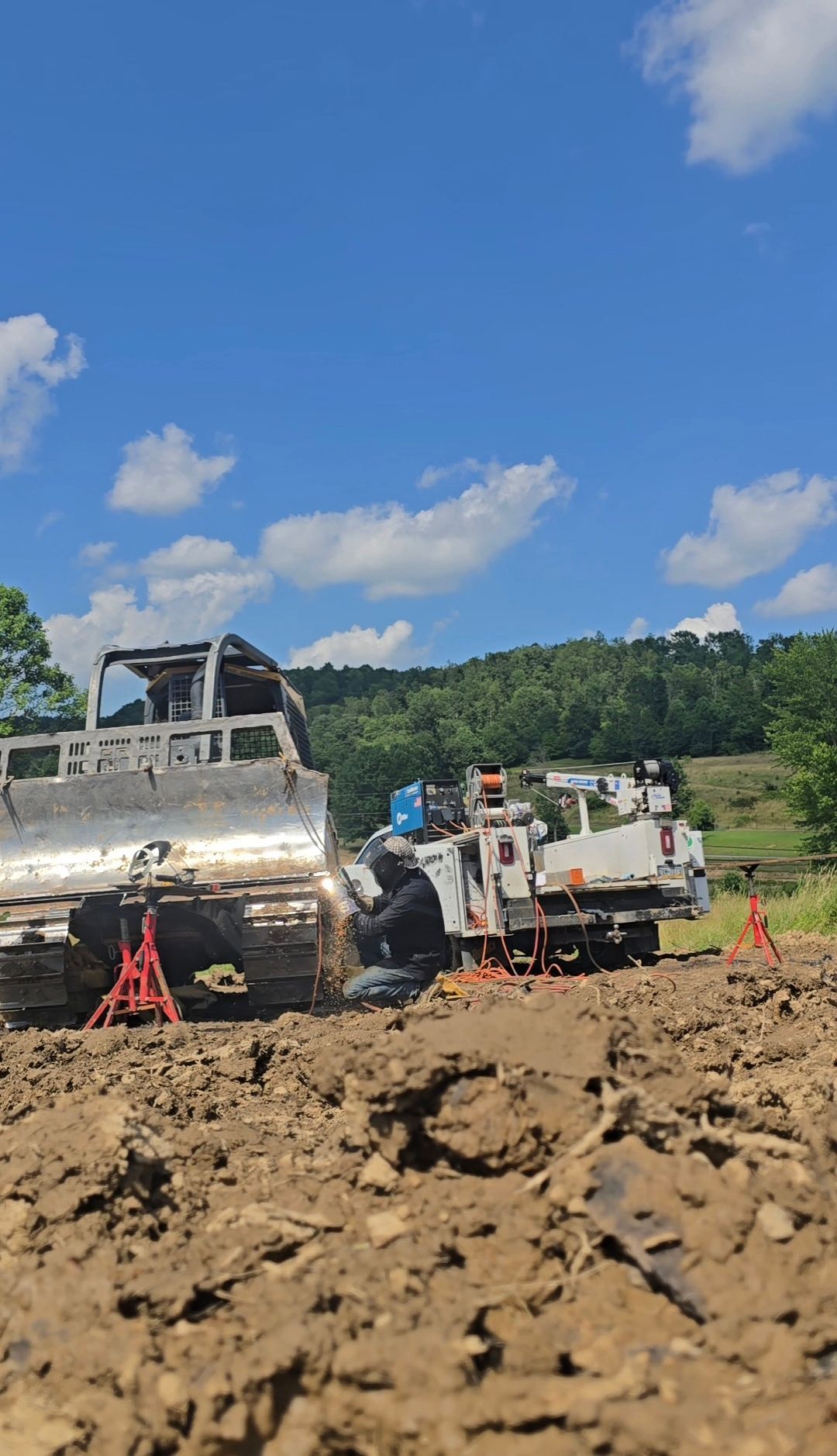 Construction trucks on a muddy site under a blue sky, with one white utility truck and a damaged vehicle in view.