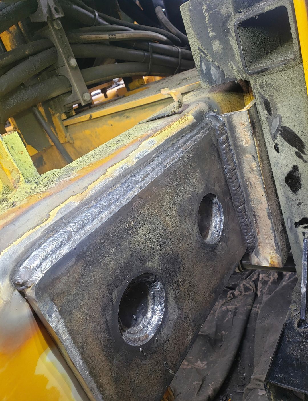 Close-up of a worn metal hinge on yellow industrial machinery with cables in the background