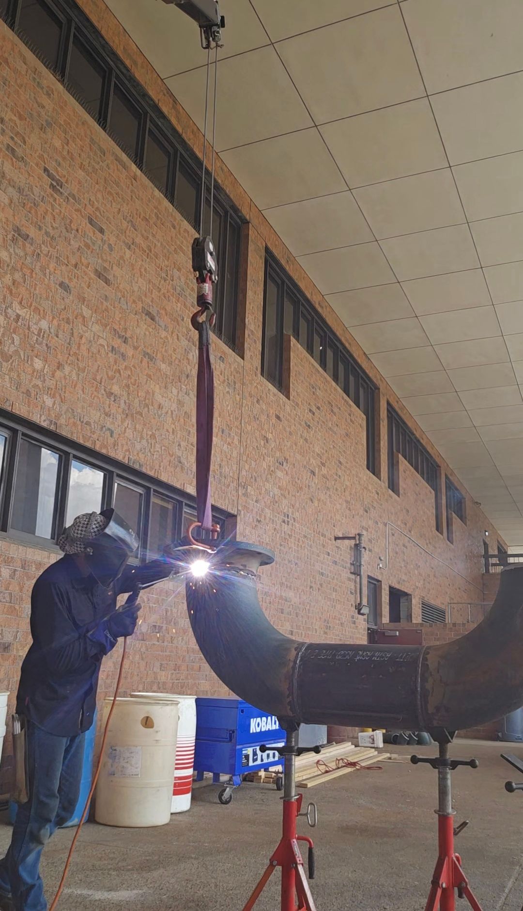 Worker welding a large metal pipe inside an industrial workshop, with bright sparks visible.