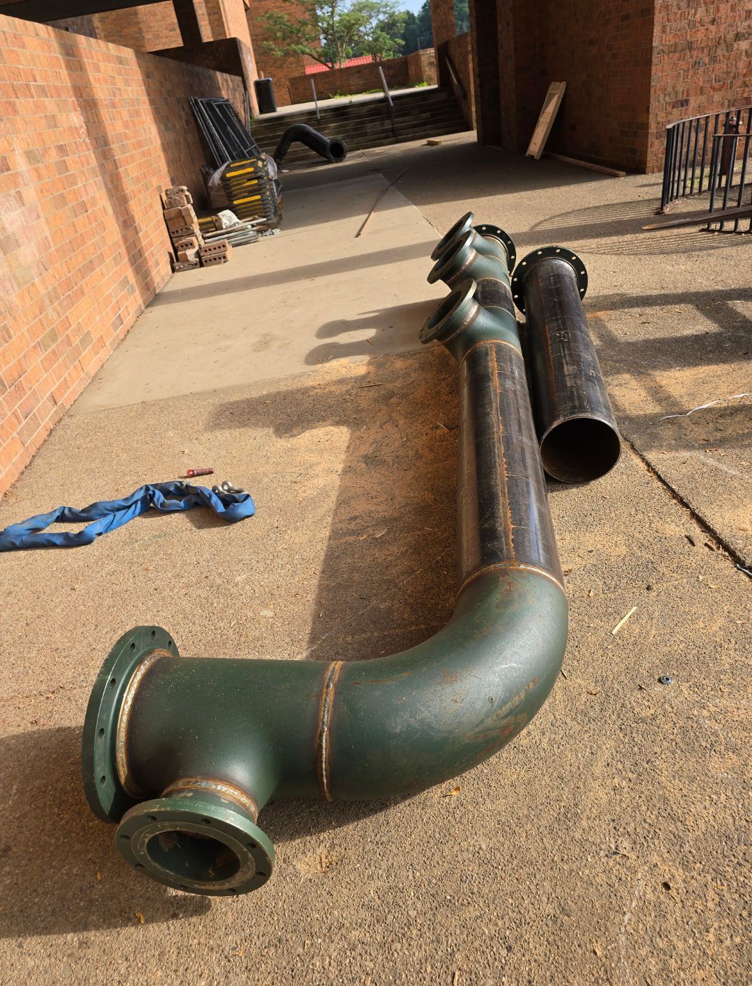 Green pipe sections on a concrete driveway beside a brick building, with tools and equipment nearby.