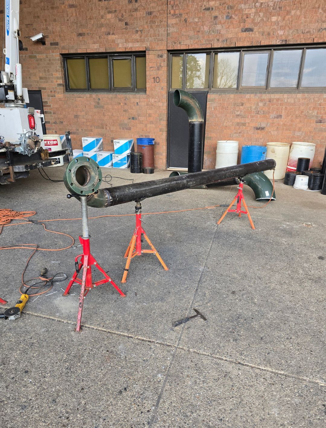 Rusty black metal pipe on red sawhorses in an industrial yard against a brick wall.