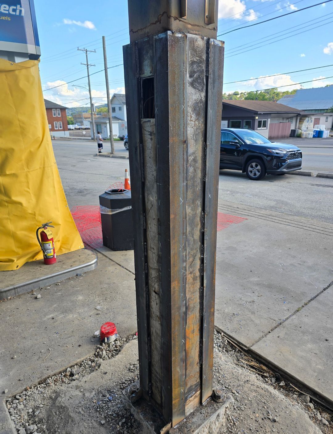 Damaged metal utility pole base with rust and broken panels on a sidewalk beside a street.