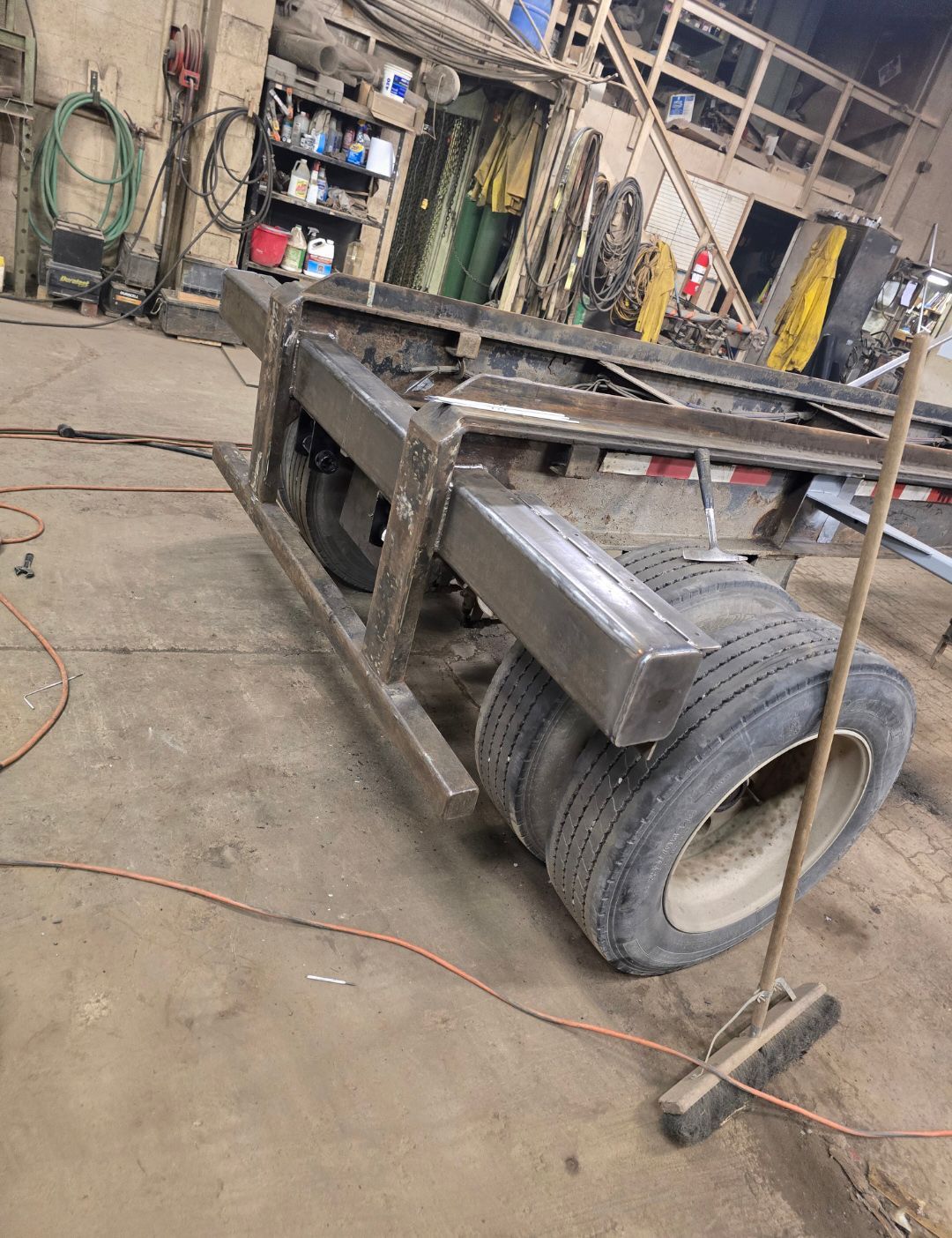 Large metal trailer frame with a tire in a workshop, viewed at an angle on a concrete floor.