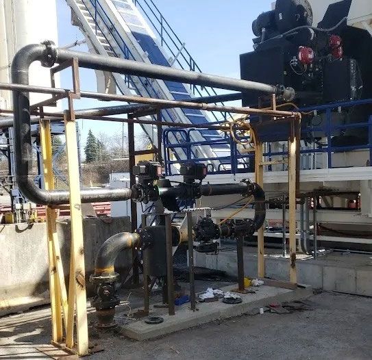 Industrial site with black pipes, yellow metal supports, and valves installed on a concrete pad near large machinery.