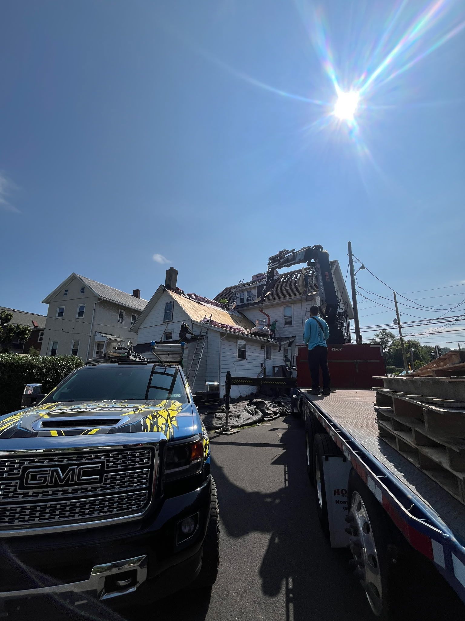Truck parked in front of a house with roof damage. Bright sunlight. Man standing nearby.
