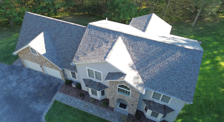 Aerial view of a two-story house with a gray roof and light-colored siding, surrounded by green grass and trees.