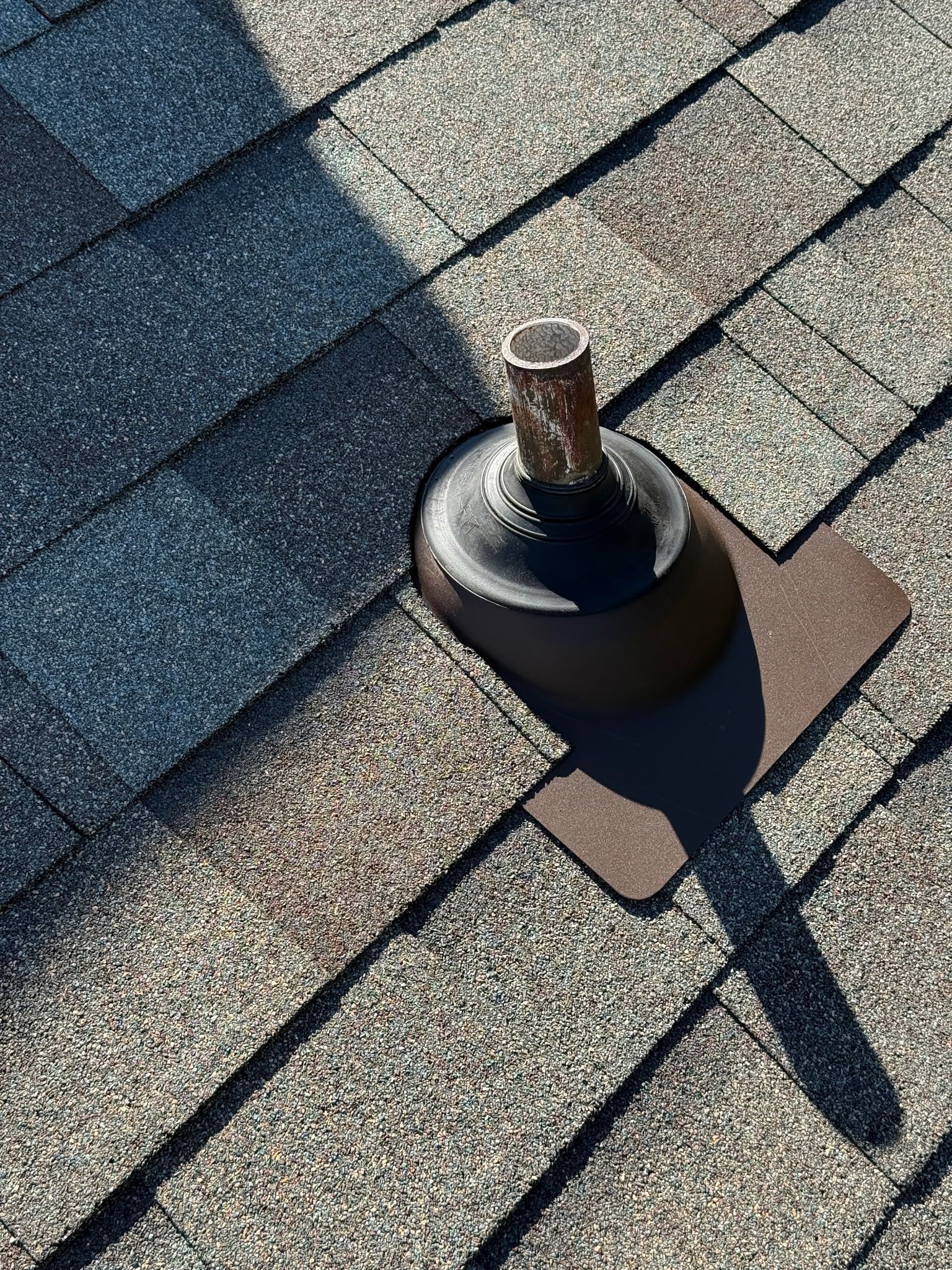A roof vent pipe protruding from gray asphalt shingles with brown base flashing.