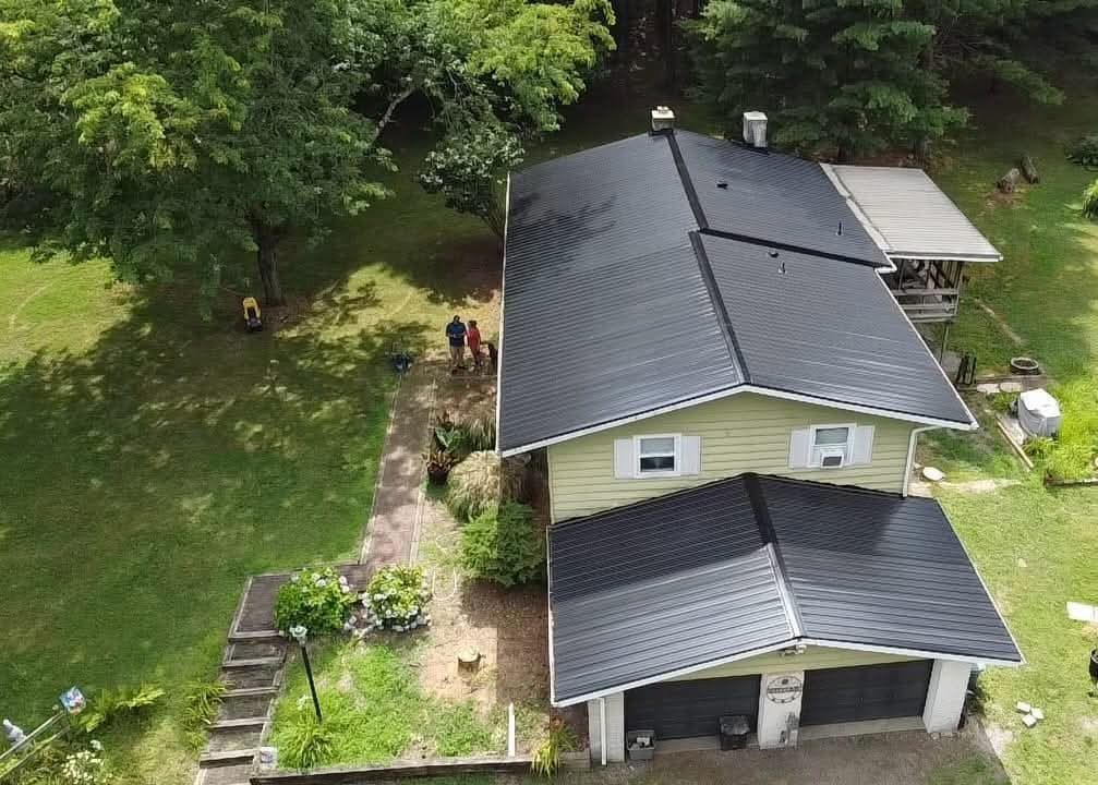 Overhead view of a two-story house with a black roof and attached garage. Green lawn with trees and people.