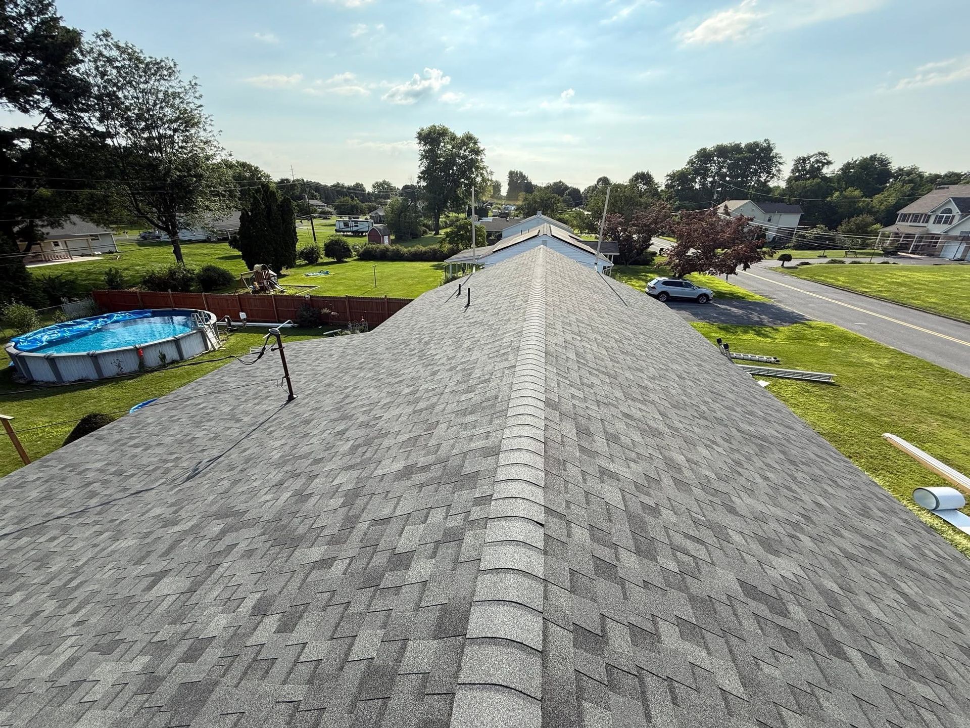 View of a residential roof with gray shingles, surrounded by greenery and homes, on a sunny day.