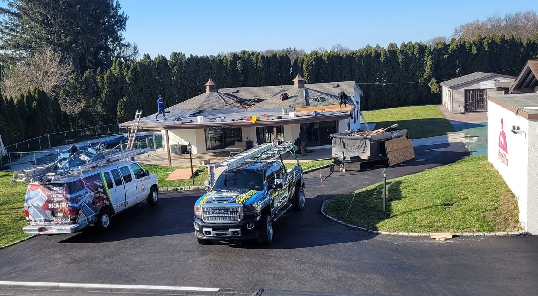 Roofing crew working on a house; trucks, materials, and ladders present.