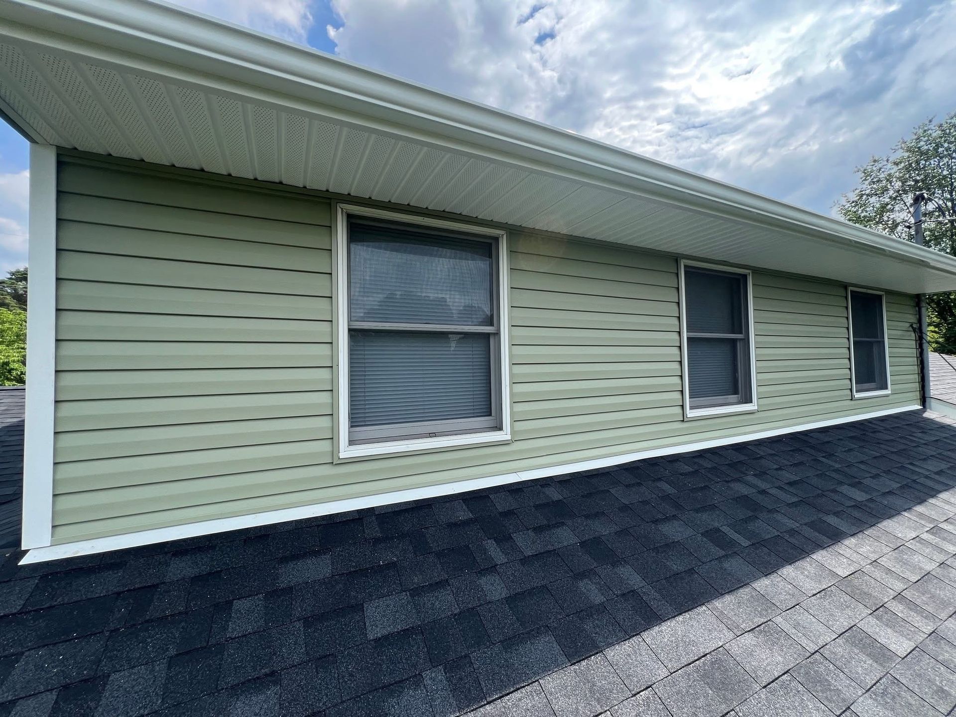 Side of a house with green siding, three windows, and a dark roof under a cloudy sky.