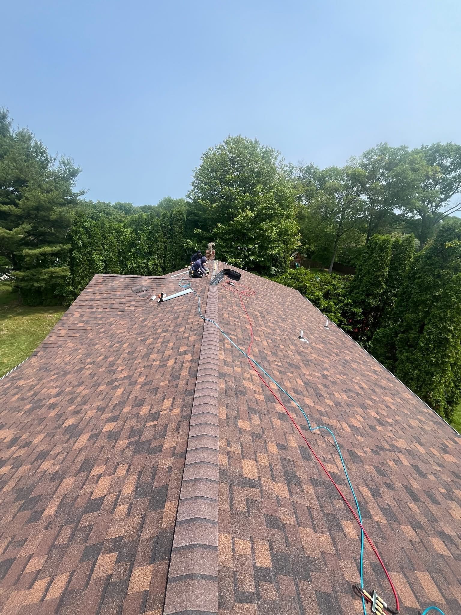 Roofers working on a brown shingled roof with trees in the background under a blue sky.