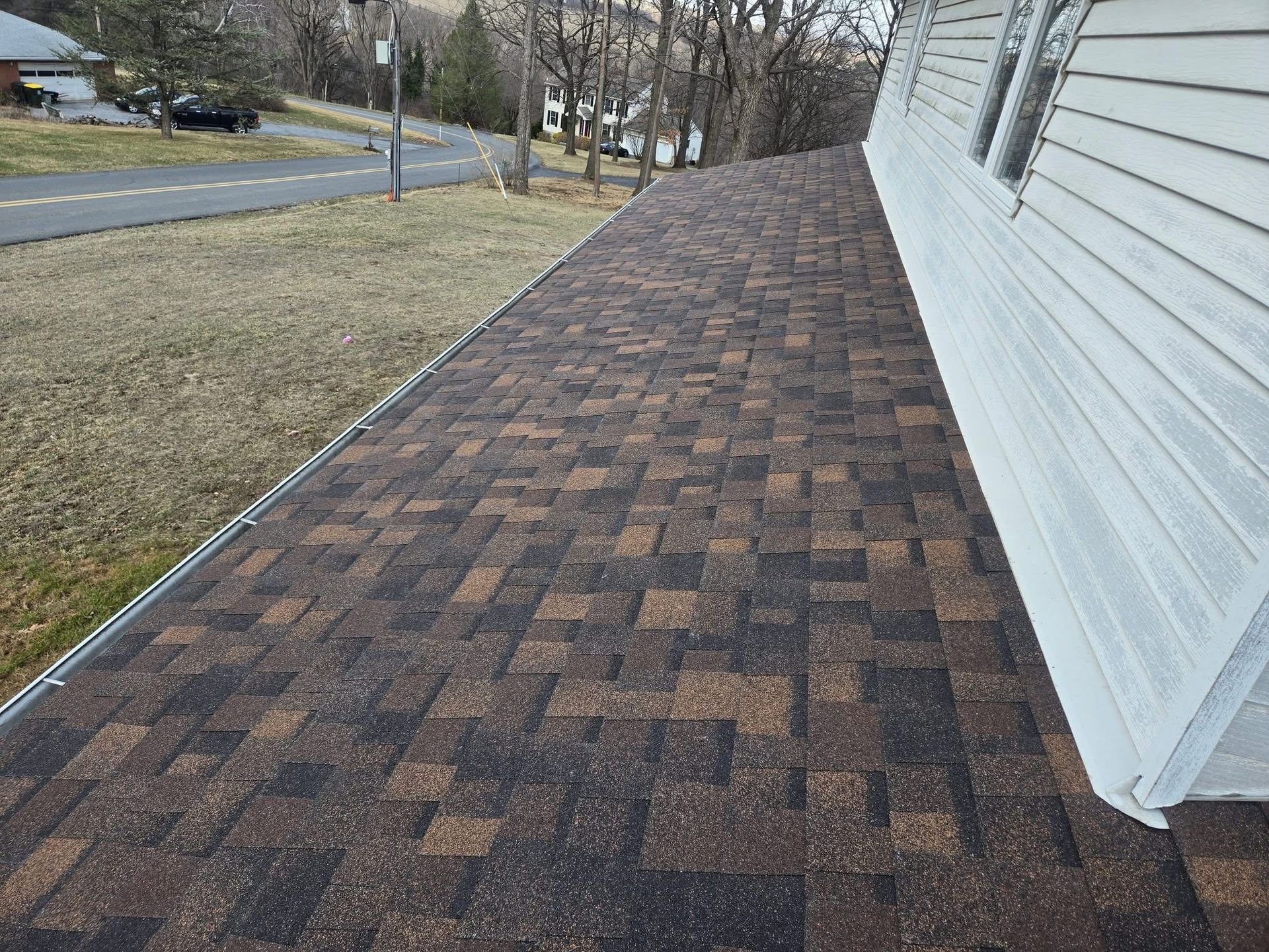 Brown and black shingle roof with silver trim, adjacent to a white-sided house and a road with a few parked cars.