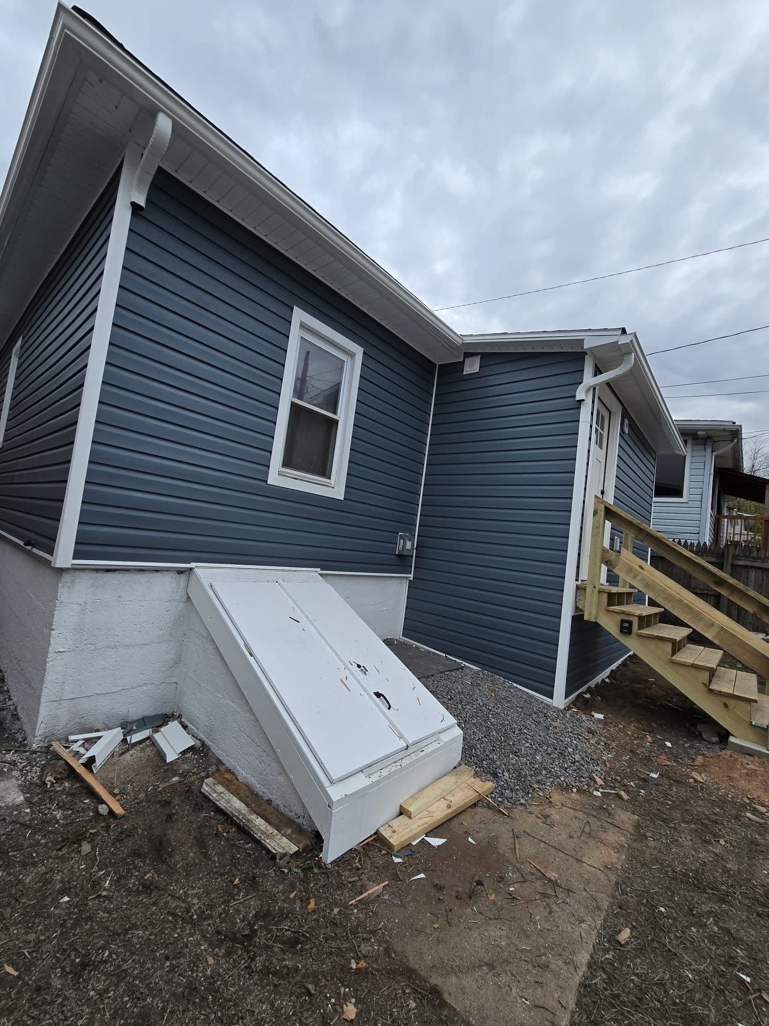 Blue-sided building with white trim, a basement entrance, and wooden stairs against a cloudy sky.